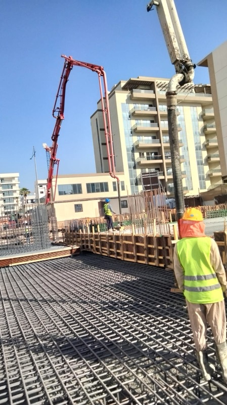 Worker in hard hat and high-visibility vest standing on a raft reinforcement mat while a concrete pump boom is positioned